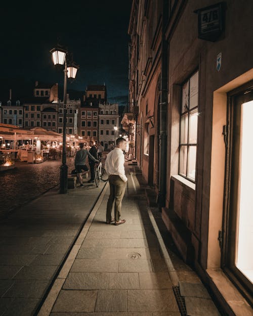 Man in White Shirt and Brown Pants Walking on Sidewalk during Night Time