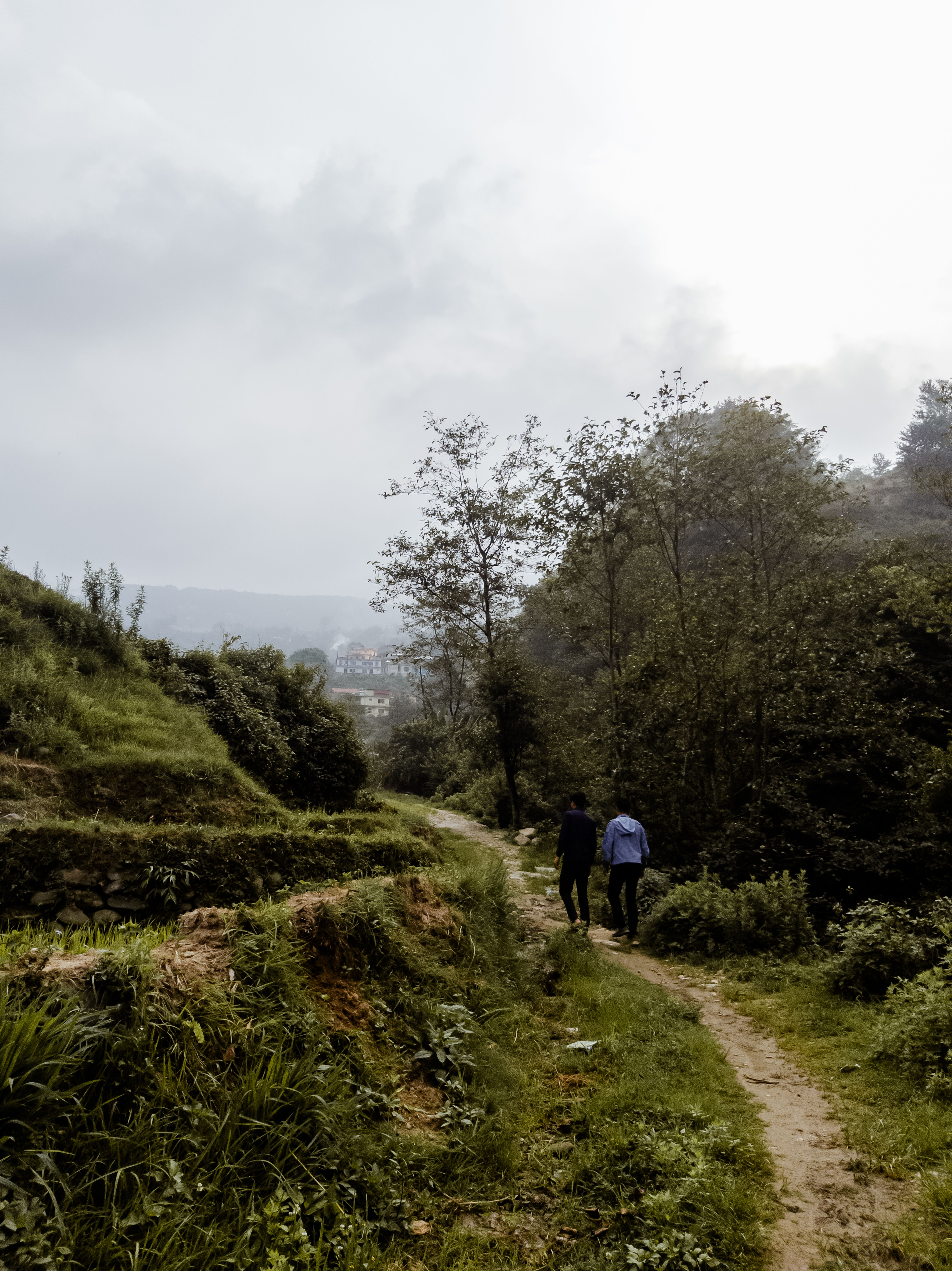 Two hikers walking a dirt path between terraced fields on a cloudy day near Kathmandu