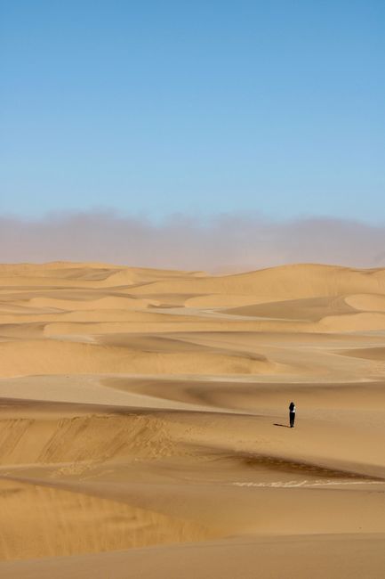 A solitary figure walking across the expansive dunes of the Namib Desert under a clear blue sky.