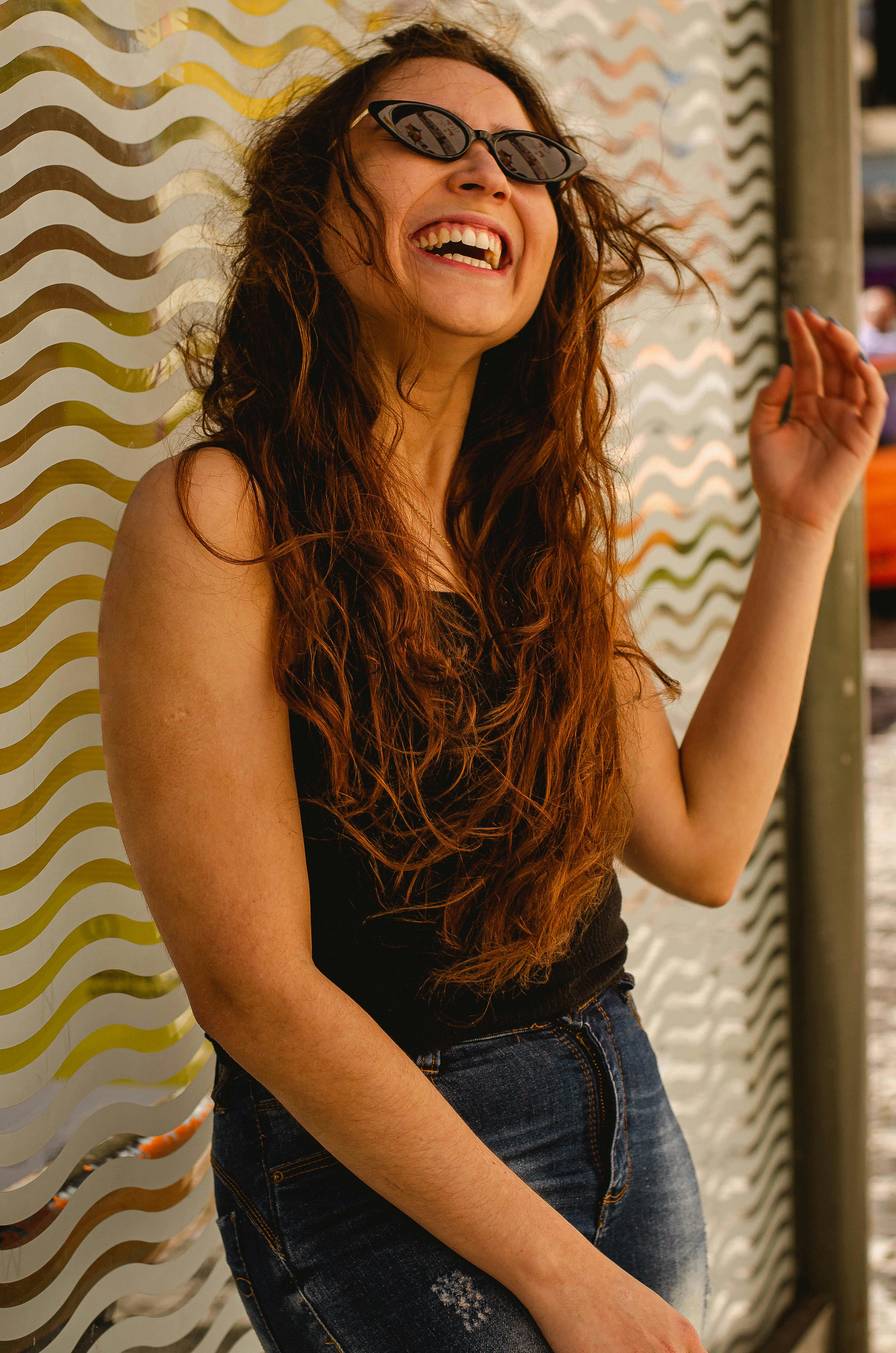 一位戴着太阳镜的快乐女人，正靠着户外一面波浪图案的墙壁开怀大笑