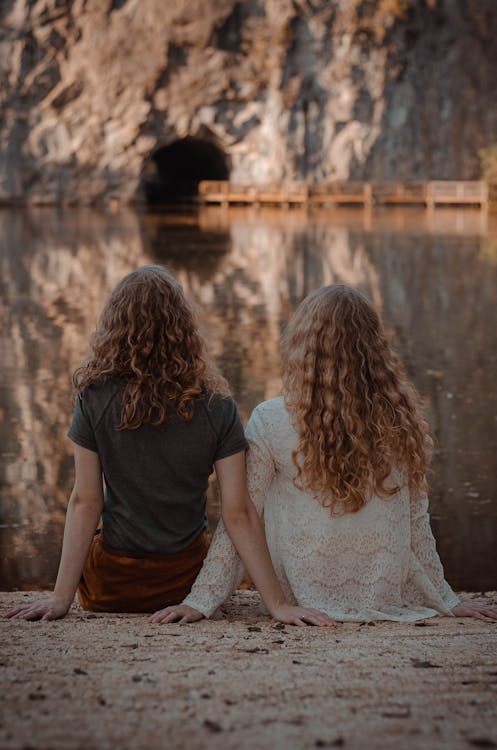 Free Photography of Two Women Sitting on Ground Facing on Body of Water Stock Photo