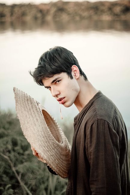 Stylish young man holding a straw hat by a serene lakeside, exuding a contemplative vibe.
