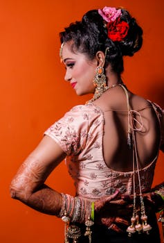 Stunning Indian bride in traditional wear with henna tattoos and jewelry on an orange backdrop.