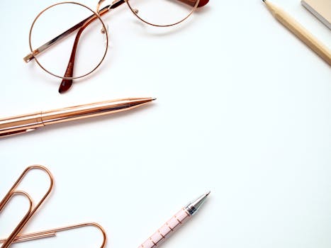 白色背景上，一支钢笔、一副眼镜和一枚回形针的特写，展现了一个时髦的极简主义书桌设置的顶视图