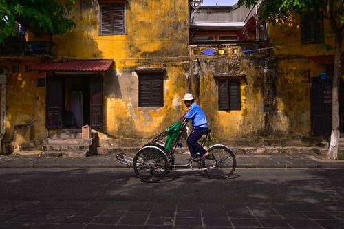 Free A man rides a tricycle past a weathered yellow wall in an urban setting. Stock Photo