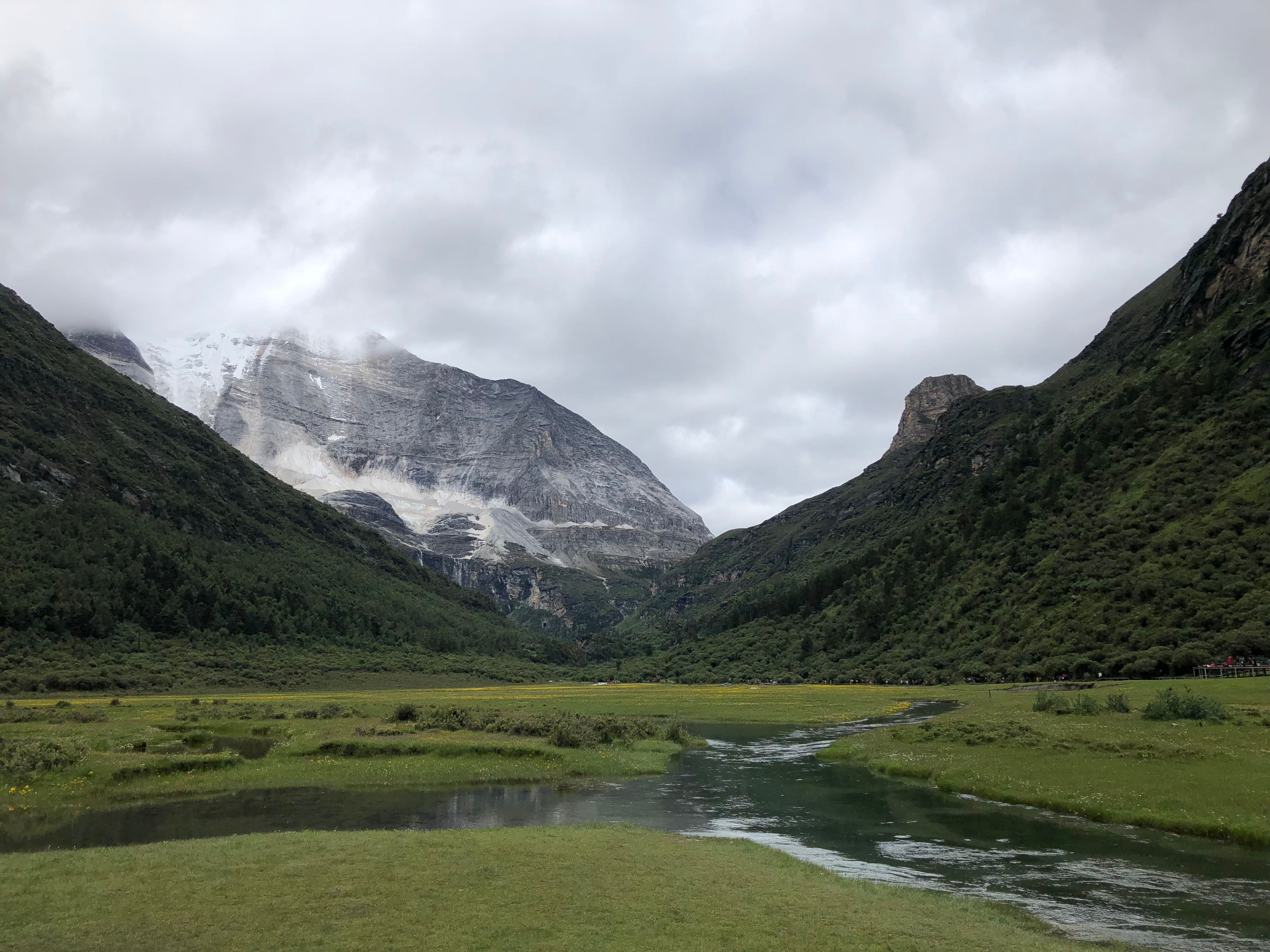 Serene valley with flowing river and majestic mountain backdrop under cloudy sky.