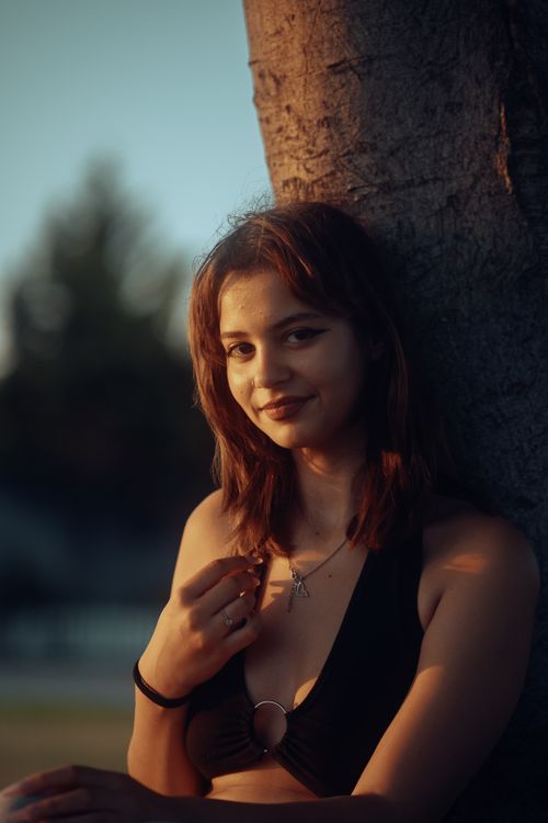Free A young woman with brown hair poses outdoors, leaning against a tree. Stock Photo