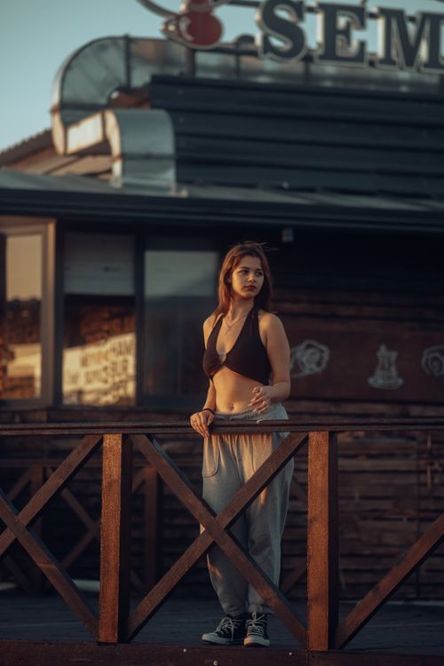 Free Stylish portrait of a young woman in a bikini top, standing near a fence in an urban setting at sunset. Stock Photo