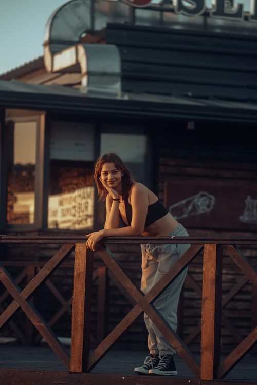 Free A smiling woman leans on a wooden fence in a casual outdoor setting, reflecting urban fashion style. Stock Photo