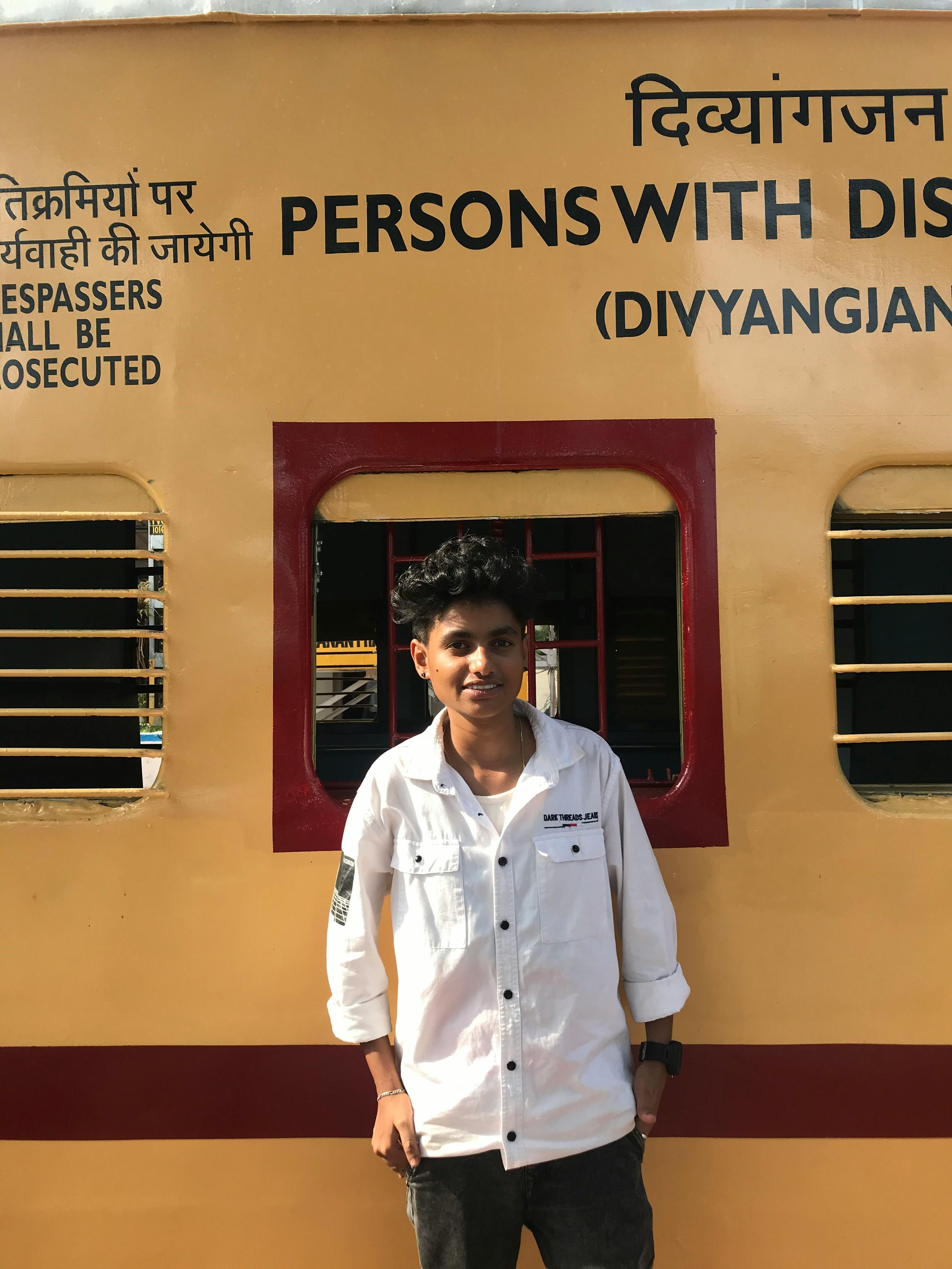 A young man stands smiling in front of a yellow train carriage with signage for persons with disabilities.