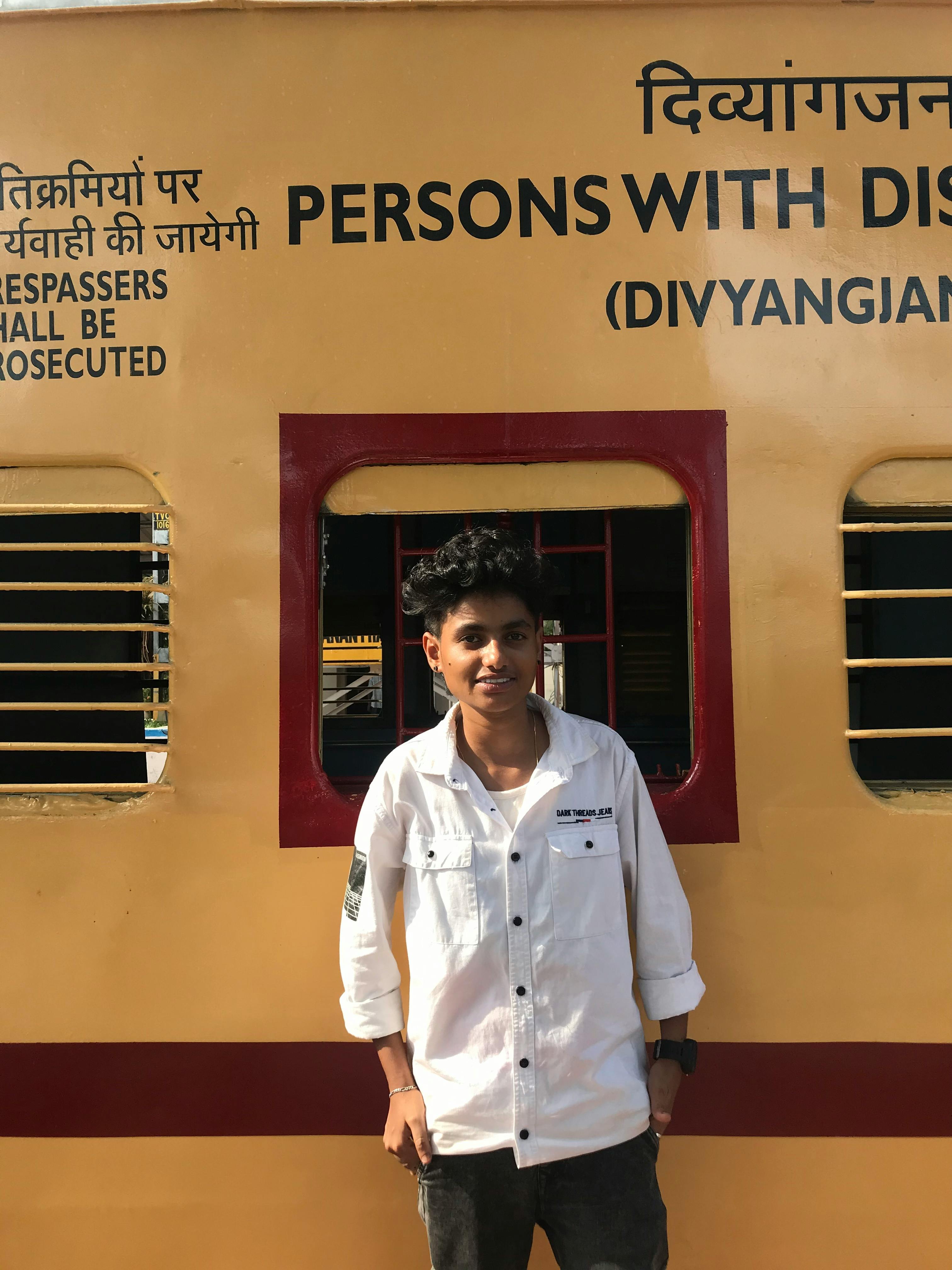 A teenager in a white shirt stands by a train carriage with prominent signage in a sunny setting.
