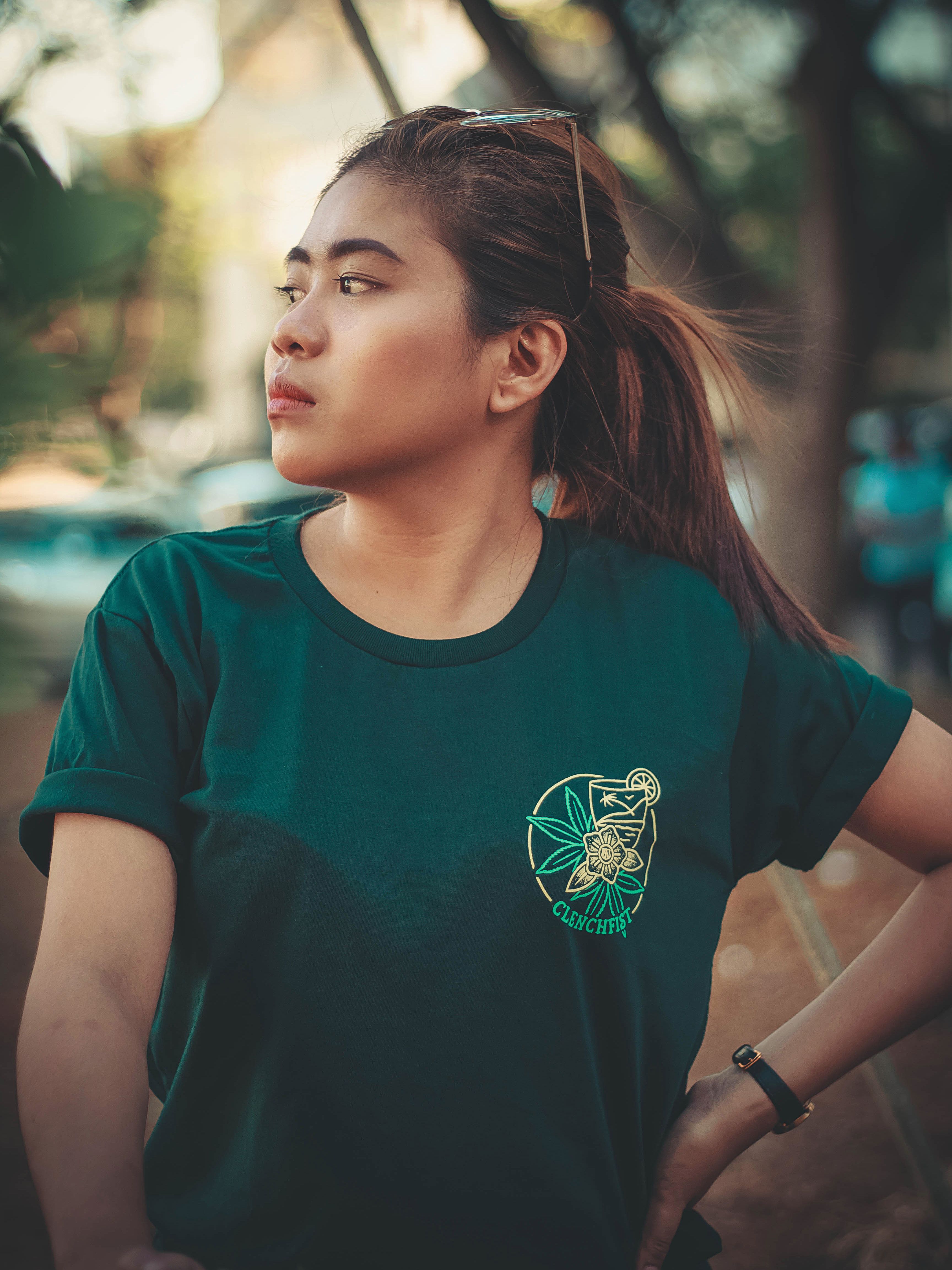 A woman wearing a green T-shirt stands outdoors, gazing thoughtfully to the side.