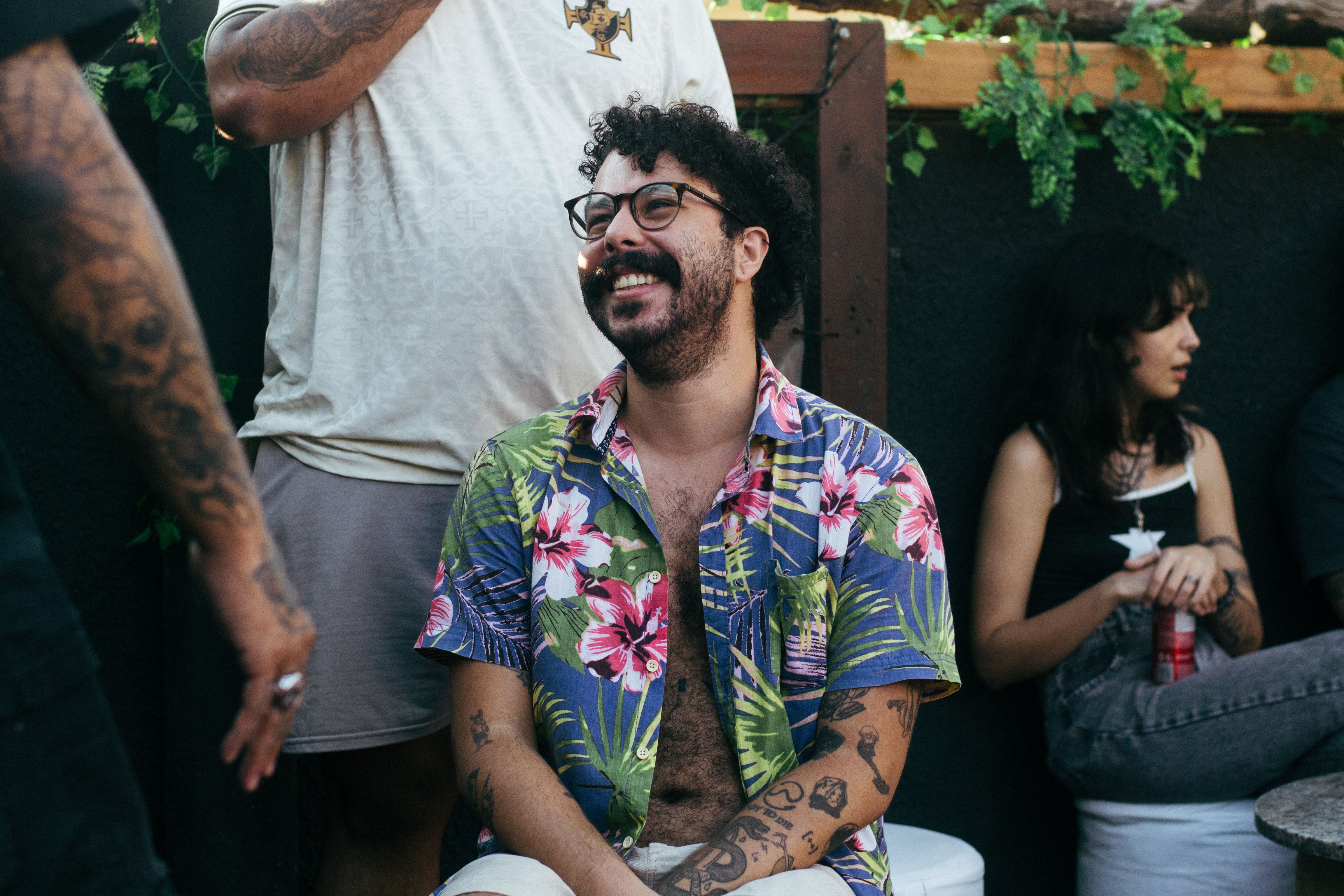 A cheerful man with tattoos and curly hair sitting outdoors, enjoying a casual moment.