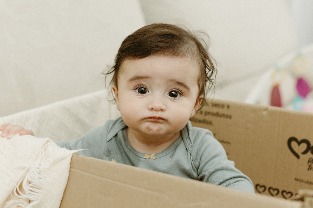 cute baby sitting in a cardboard box with a confused expression indoor