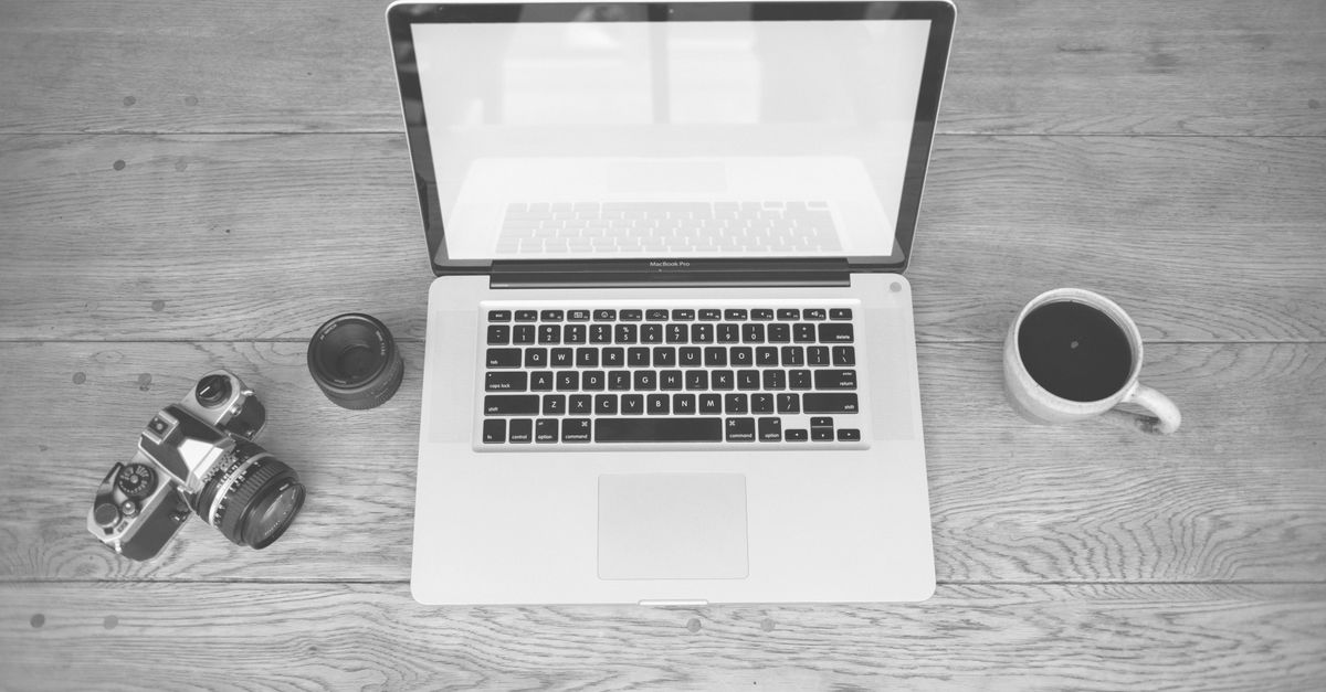 Monochrome image of a laptop, camera, lens, and coffee cup on a wooden desk