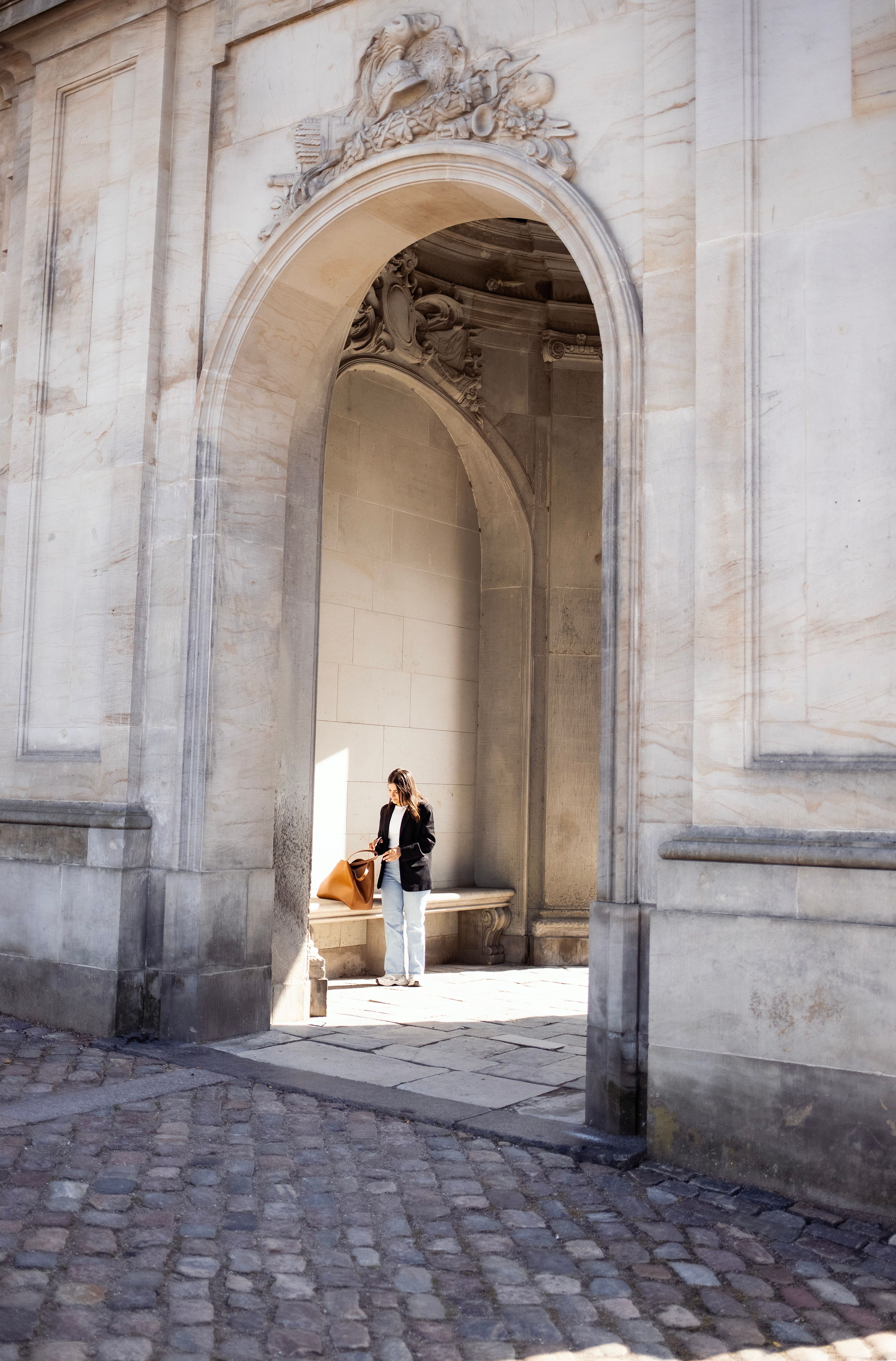 A woman stands under a historic stone archway in Copenhagen, Denmark, on a sunny day.