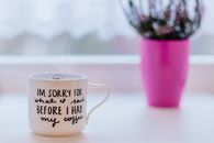 A steaming coffee mug with humorous text, set on a table near a pink plant pot indoors.