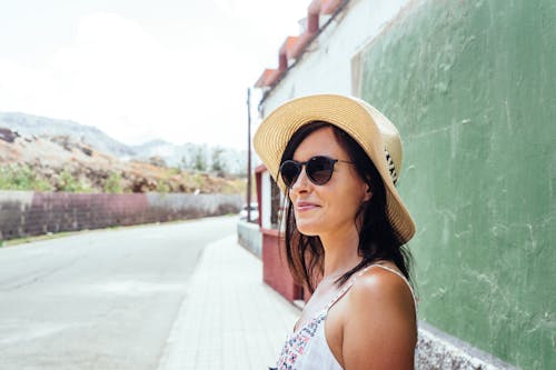 Nahaufnahmefoto Der Lächelnden Frau In Der Sonnenbrille Und Im Sonnenhut, Die Auf Bürgersteig Vor Grüner Wand Stehen