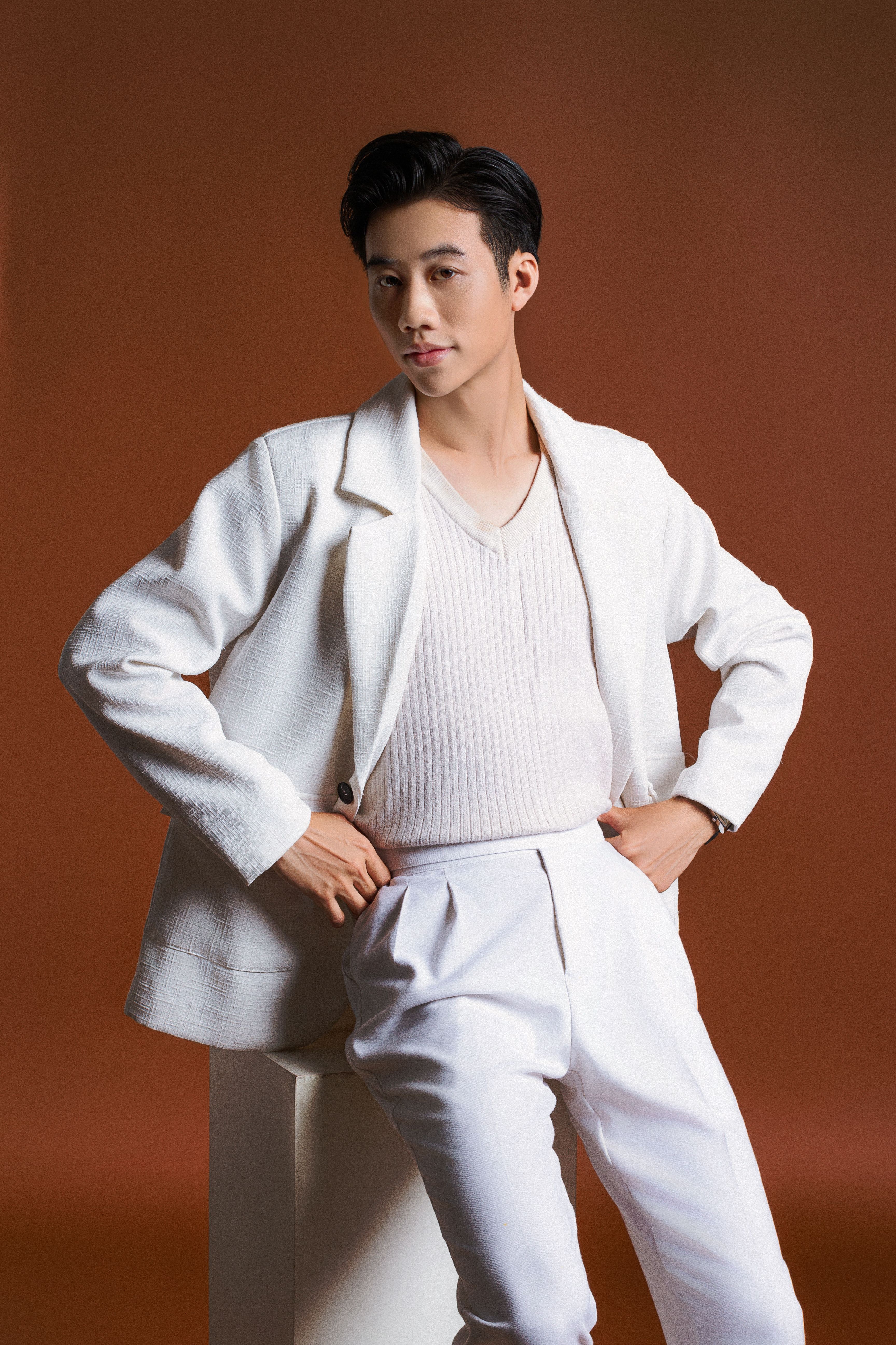Portrait of an elegant Asian man in a white suit against a brown background.
