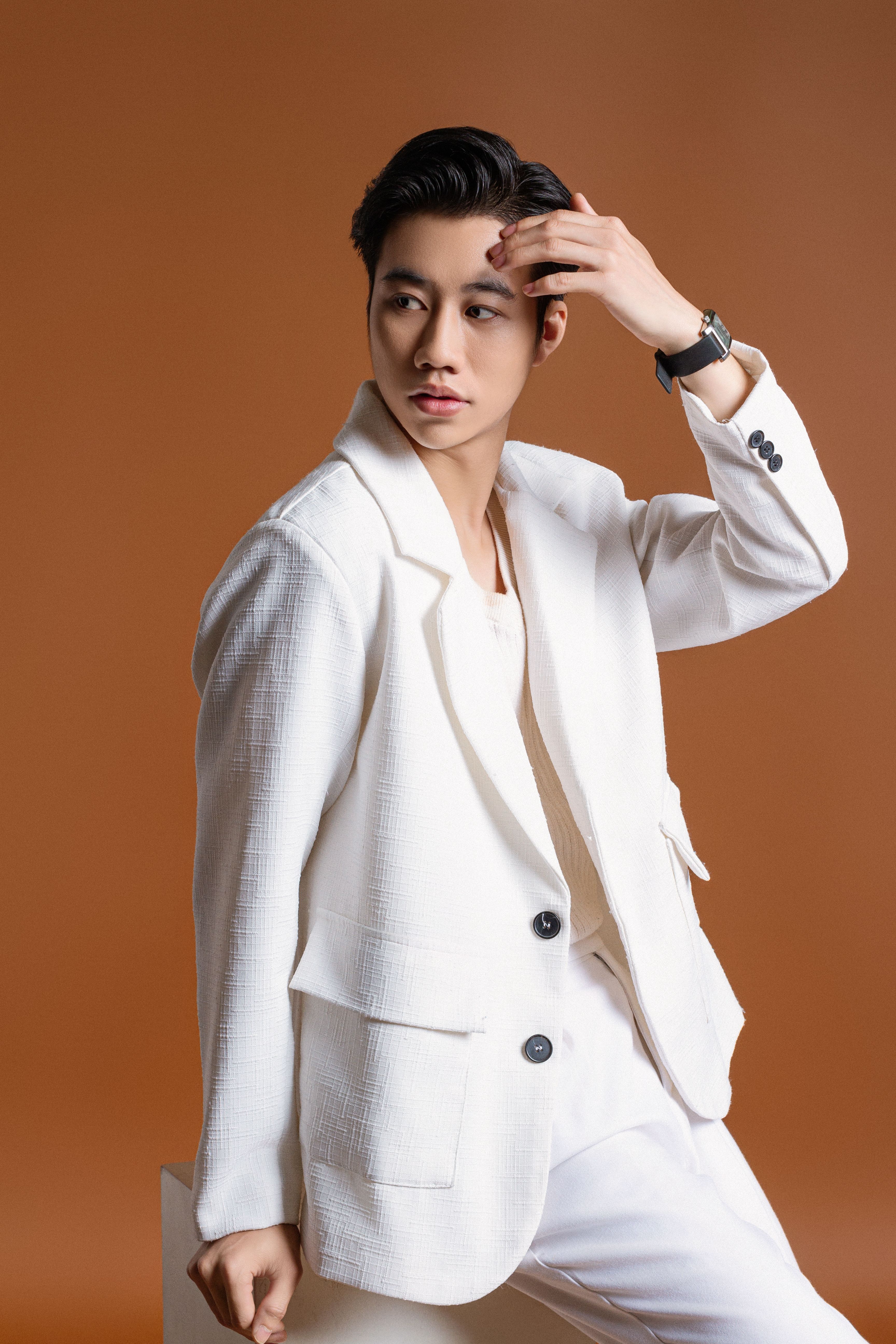 Fashion model posing in a white suit in a studio against a brown background.