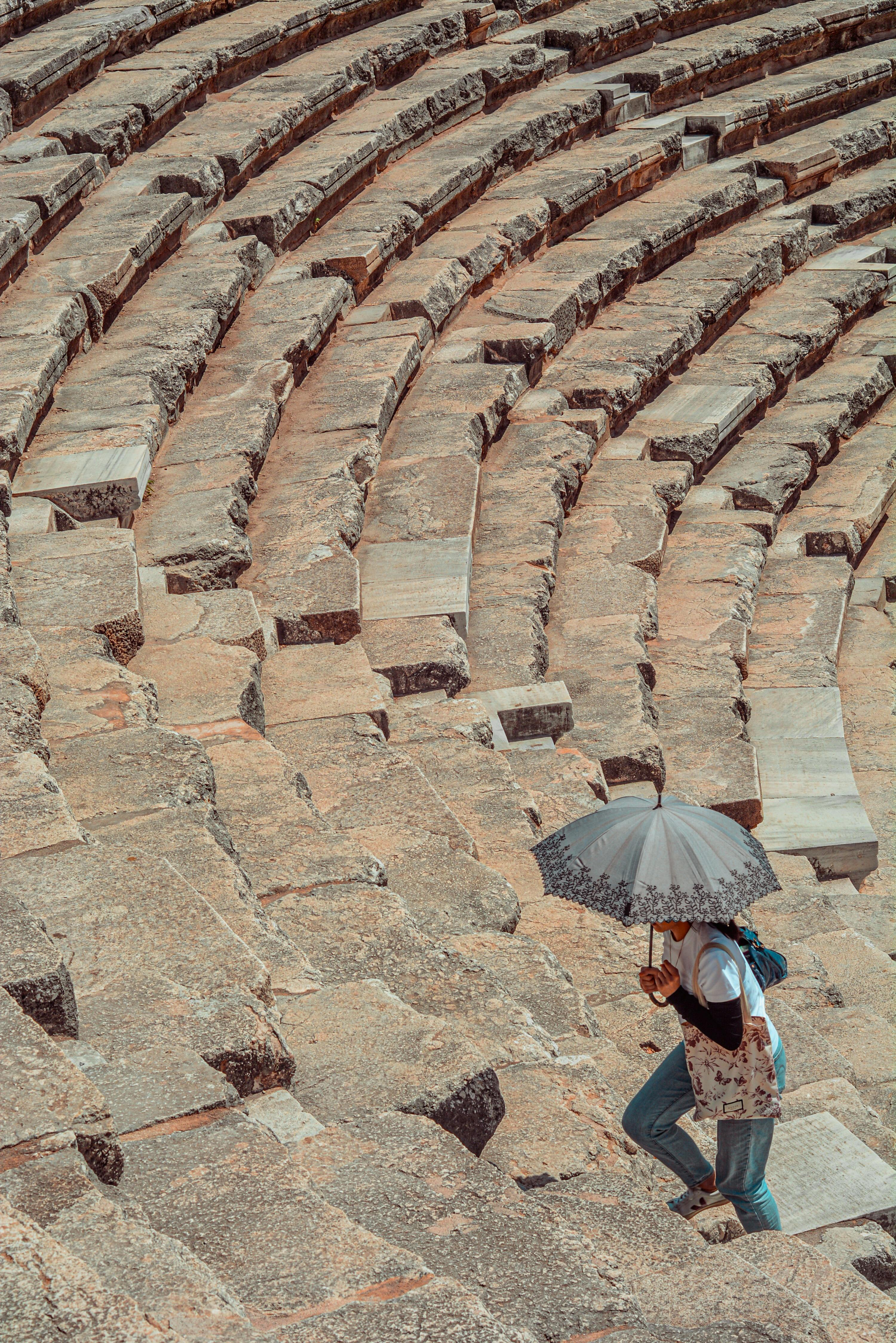 Free A person with an umbrella walks on the ancient stone steps of a theater in Antalya, Türkiye. Stock Photo