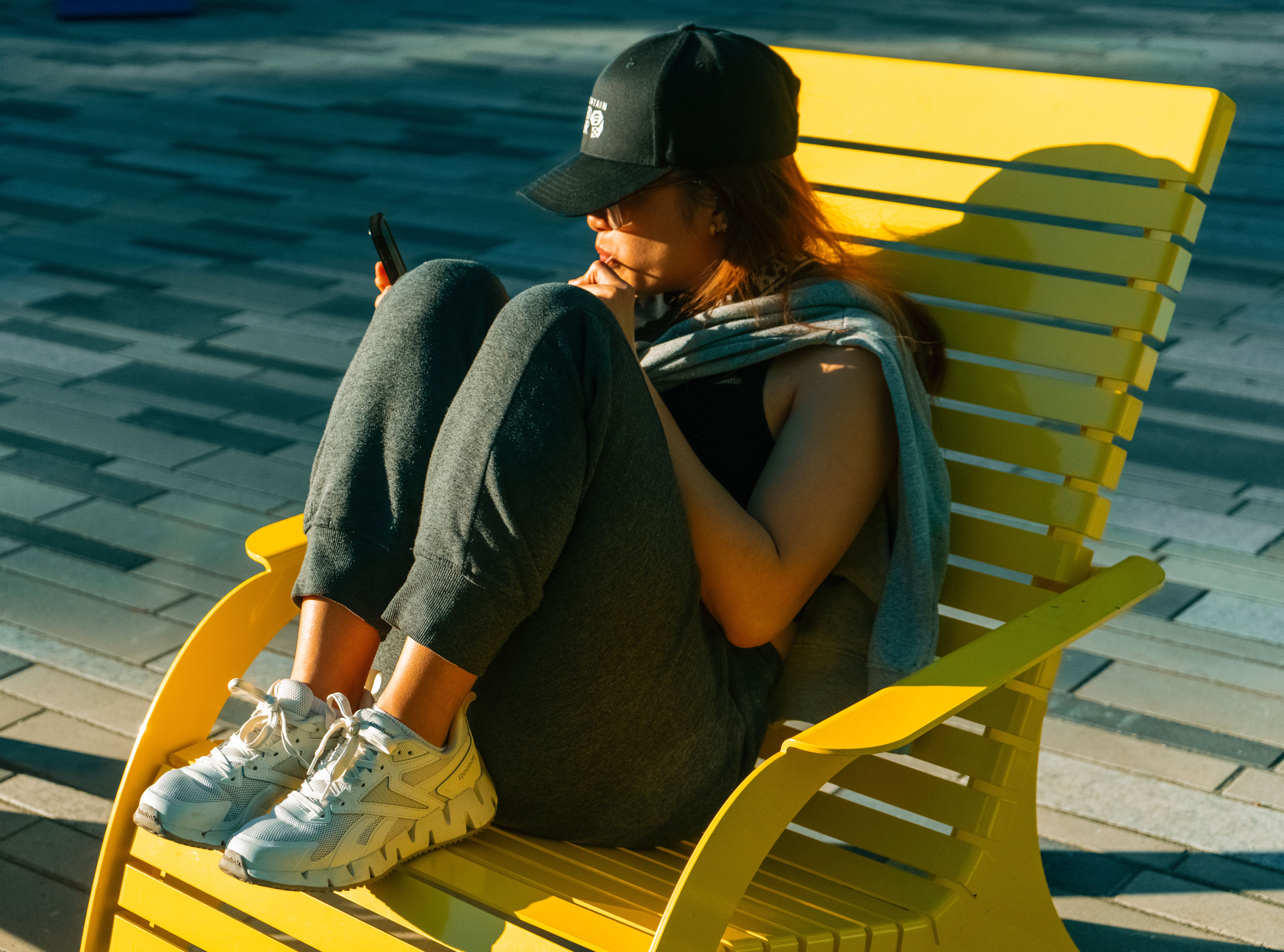 Casual woman in a cap and sneakers relaxing on a yellow chair outdoors, enjoying sunny day.