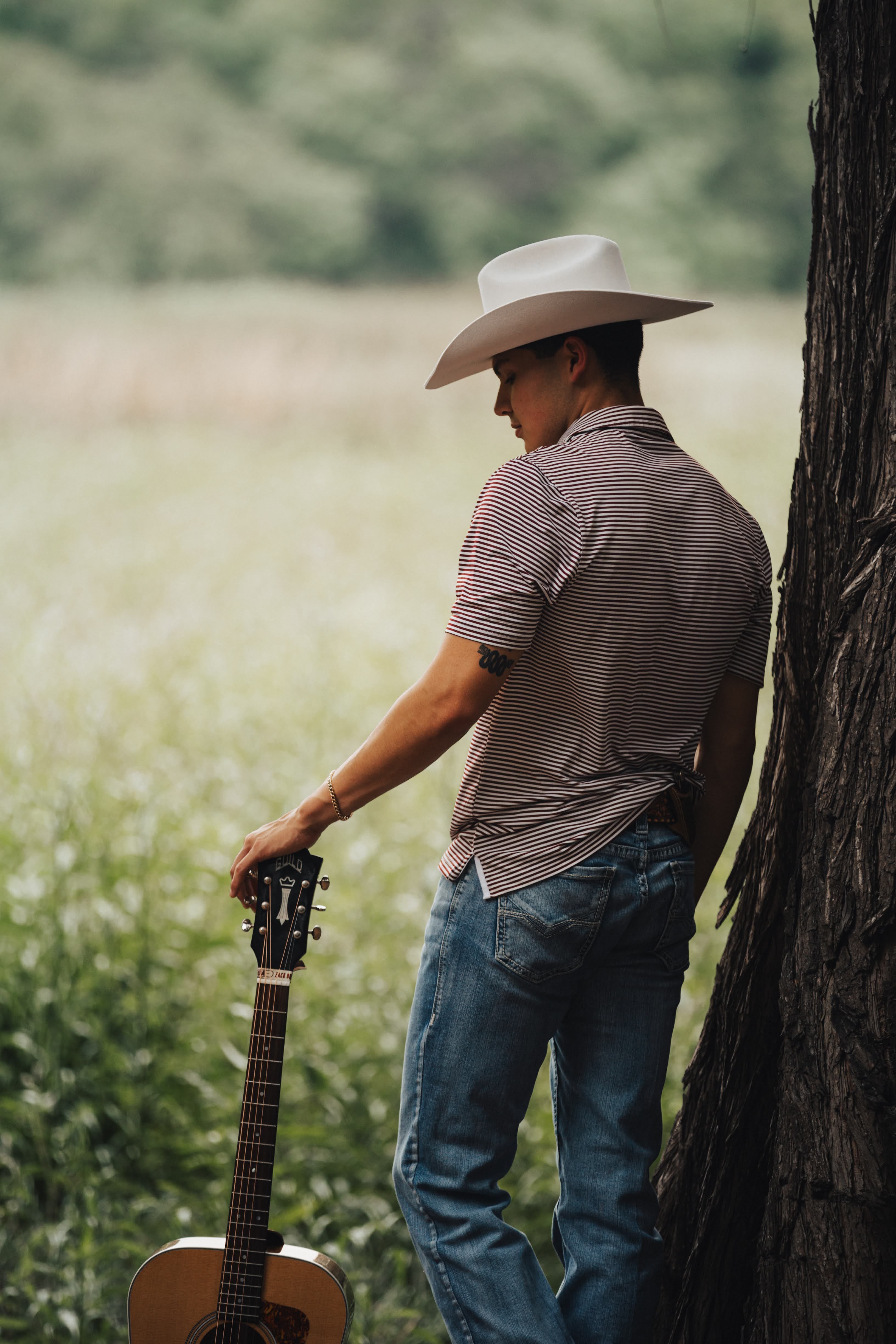 A cowboy in a hat stands by a tree, holding a guitar, immersed in nature.