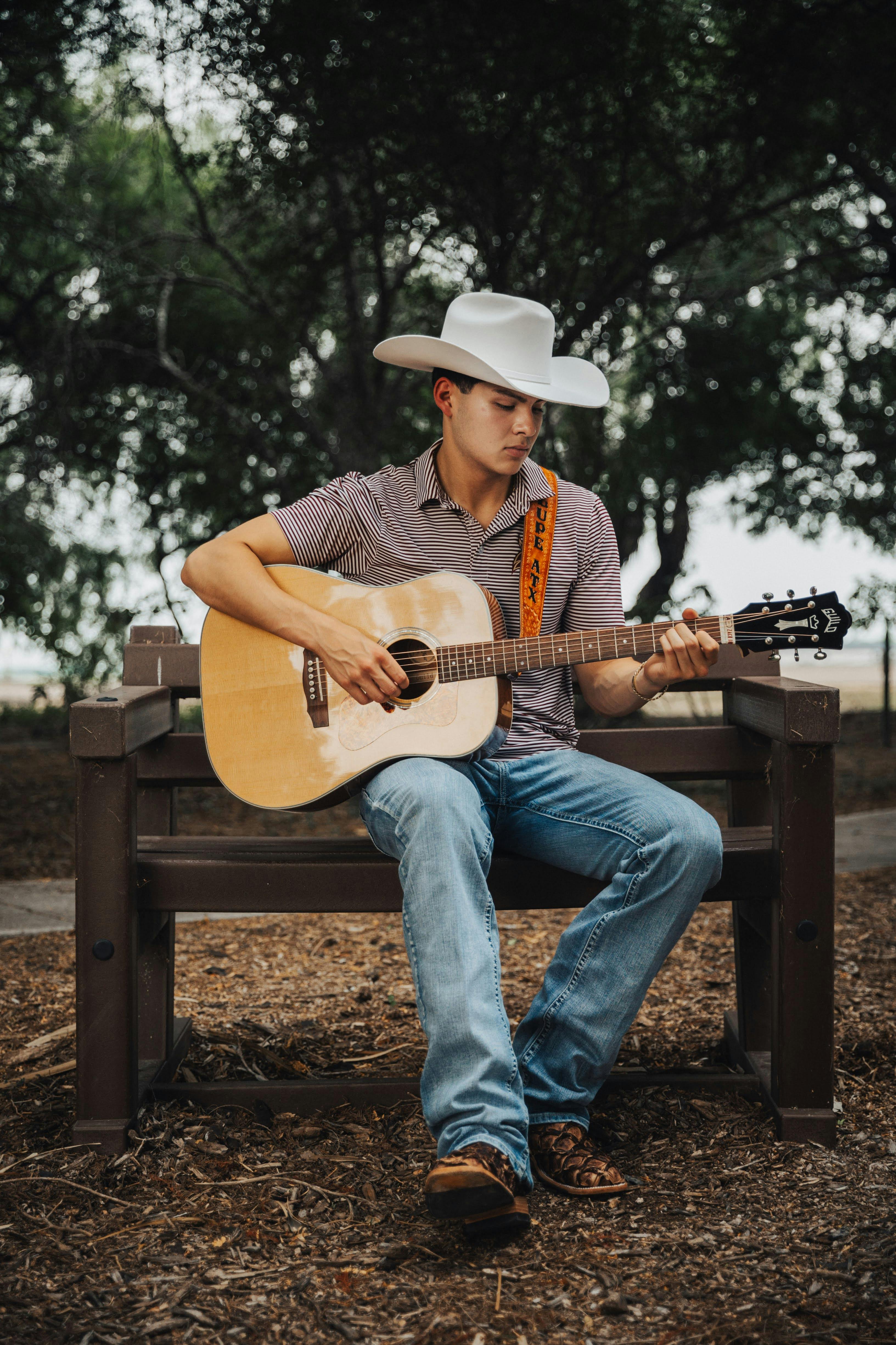 Man in cowboy hat playing acoustic guitar on park bench surrounded by trees.