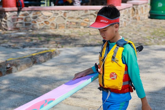 A child carries a colorful windsurf board wearing a life vest and sun visor on a sunny outdoor day.