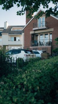A serene residential street view of a brick house with balconies and cars parked outside, surrounded by lush greenery.