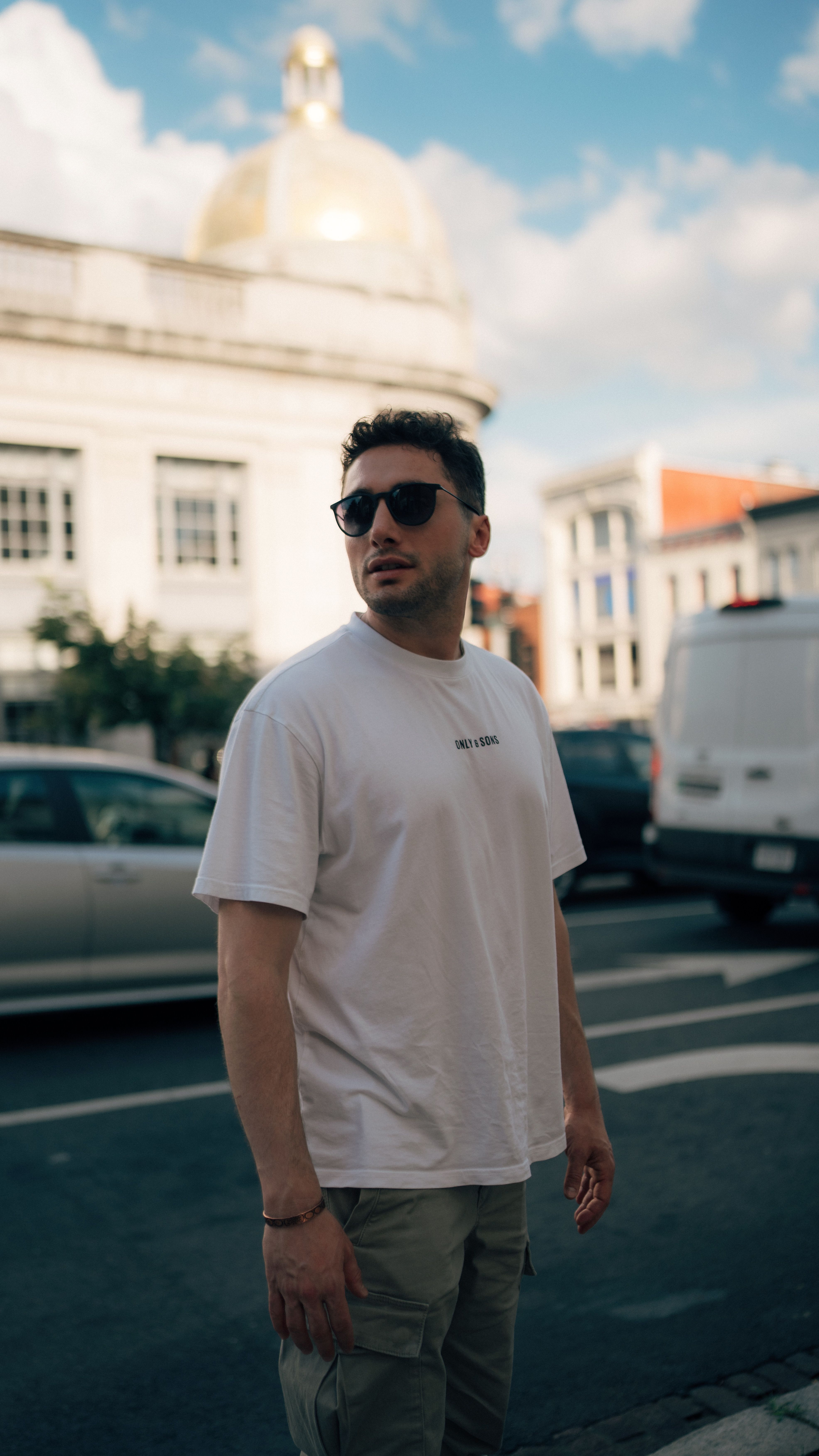 Stylish man wearing sunglasses standing on a city street with urban backdrop.