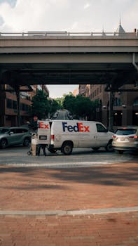 FedEx delivery van parked under an urban bridge in a busy city street scene with cars.