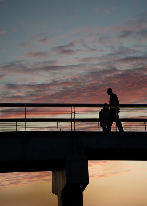 Free A man in silhouette walks along a footbridge against a vibrant sunset sky. Stock Photo