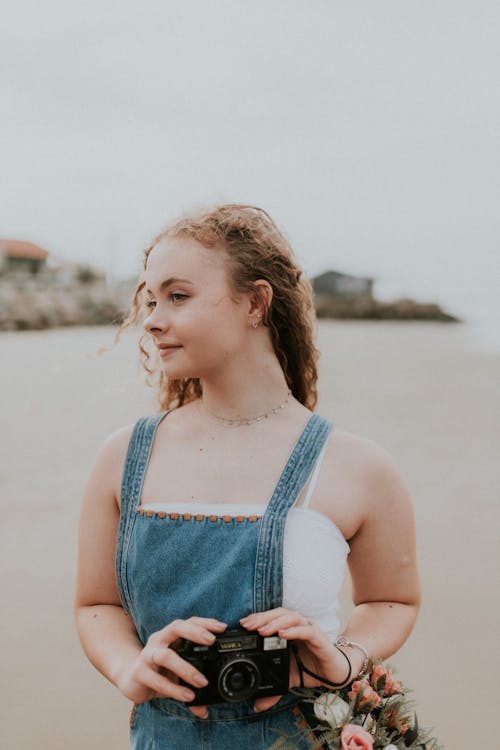 Free A girl in overalls holding a camera on the beach Stock Photo