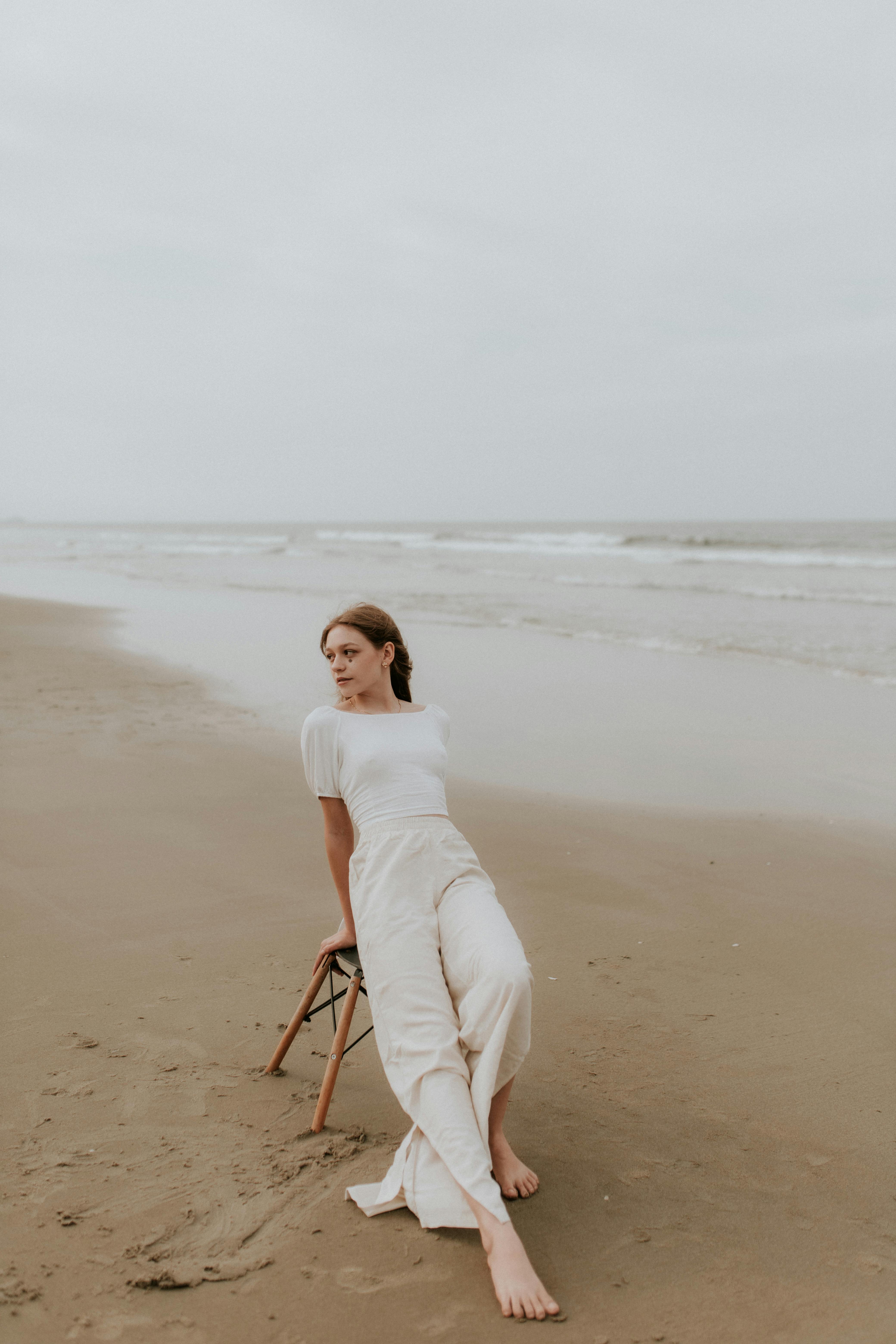 A stylish woman in neutral attire sits serenely on a sandy beach, capturing a moment of relaxation.