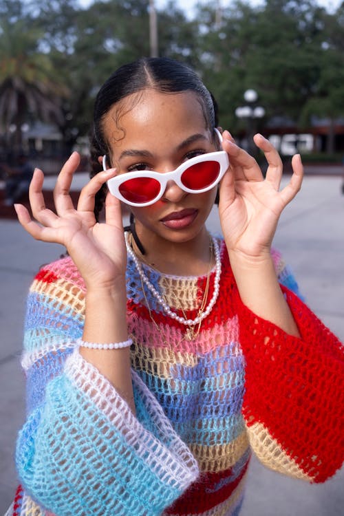 Free Stylish young woman wearing vibrant sweater and red sunglasses poses confidently outdoors. Stock Photo
