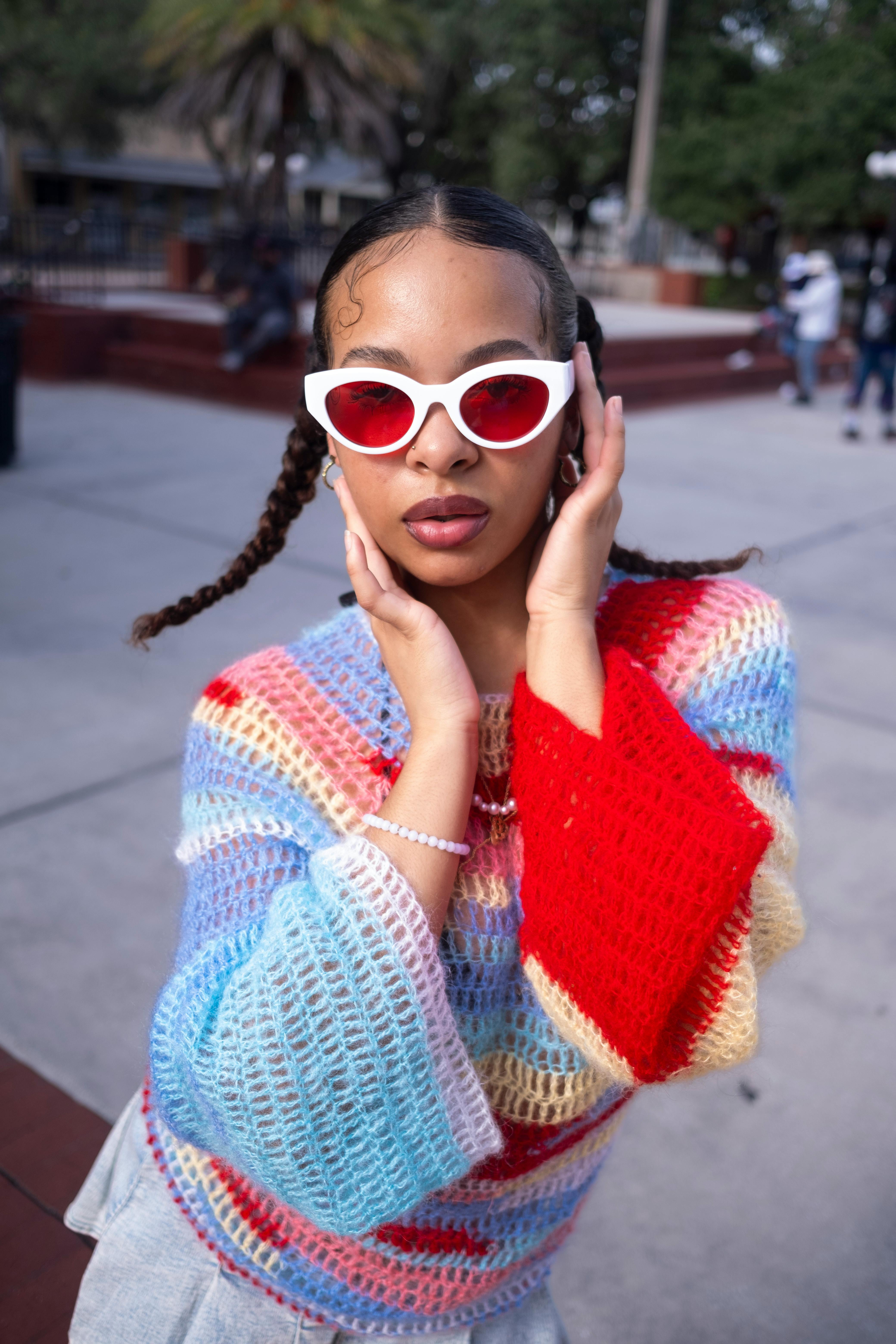 Fashionable woman posing outdoors with red sunglasses and a colorful knit sweater, exuding retro style.