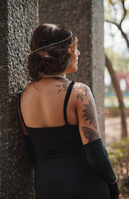 A woman with tattoos and a black dress leaning against a wall