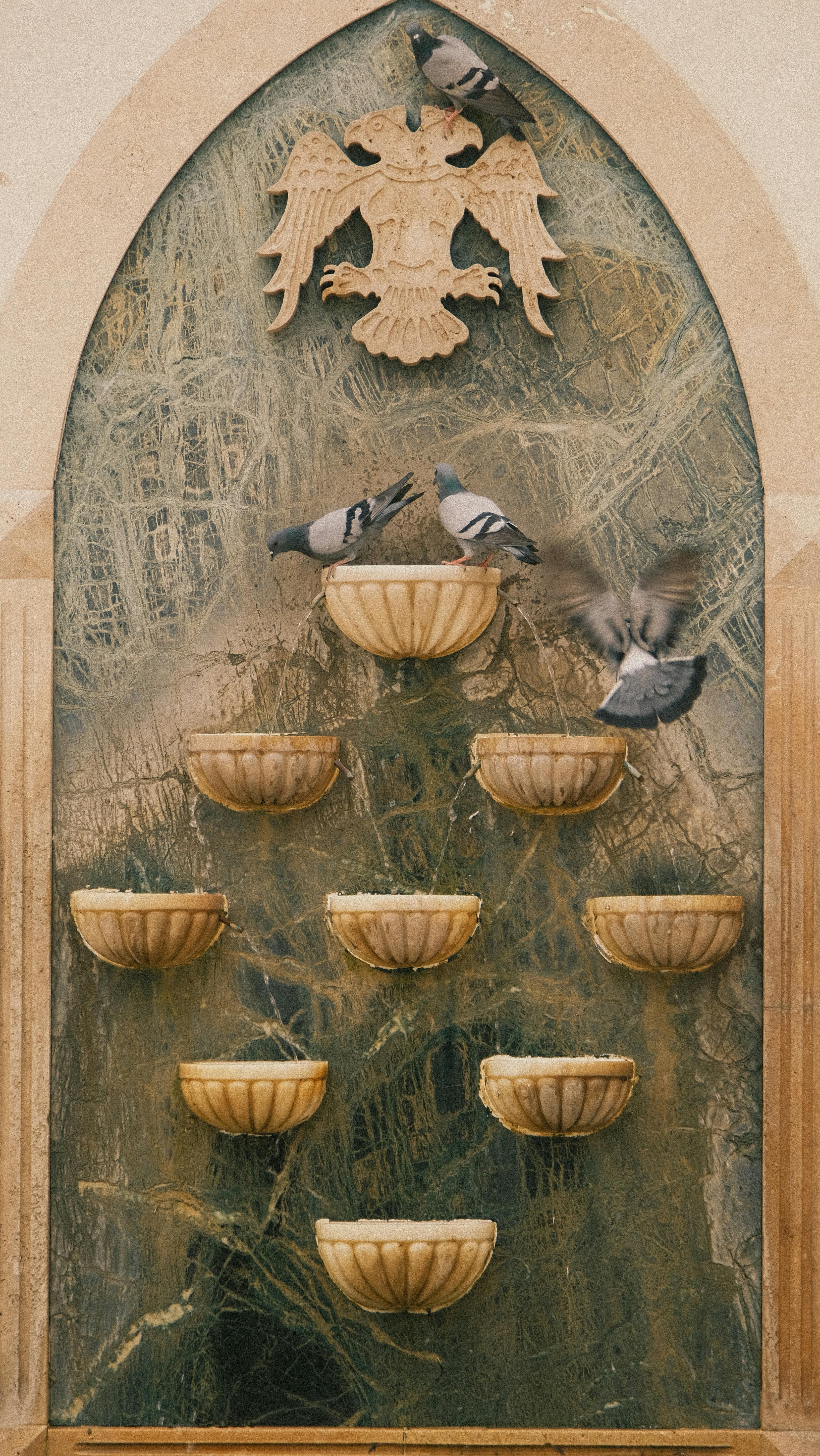A vertical stone fountain with pigeons resting amidst sculpted bowls and emblem decor.