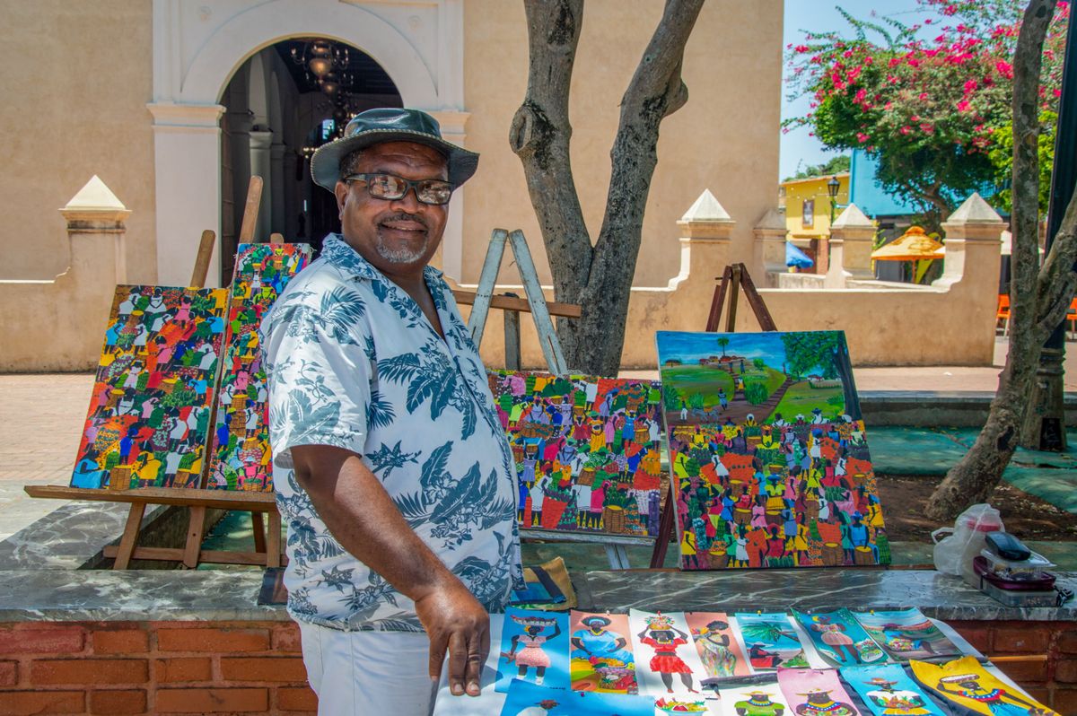 Artista local sonríe en su puesto de arte colorido en un ambiente soleado de mercado al aire libre.