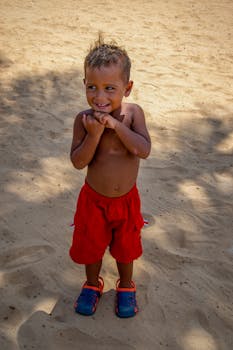 A cute child smiling in the sunlight on a sandy beach, wearing red shorts.
