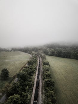 Aerial view of railroad tracks cutting through a foggy countryside landscape surrounded by lush green fields.