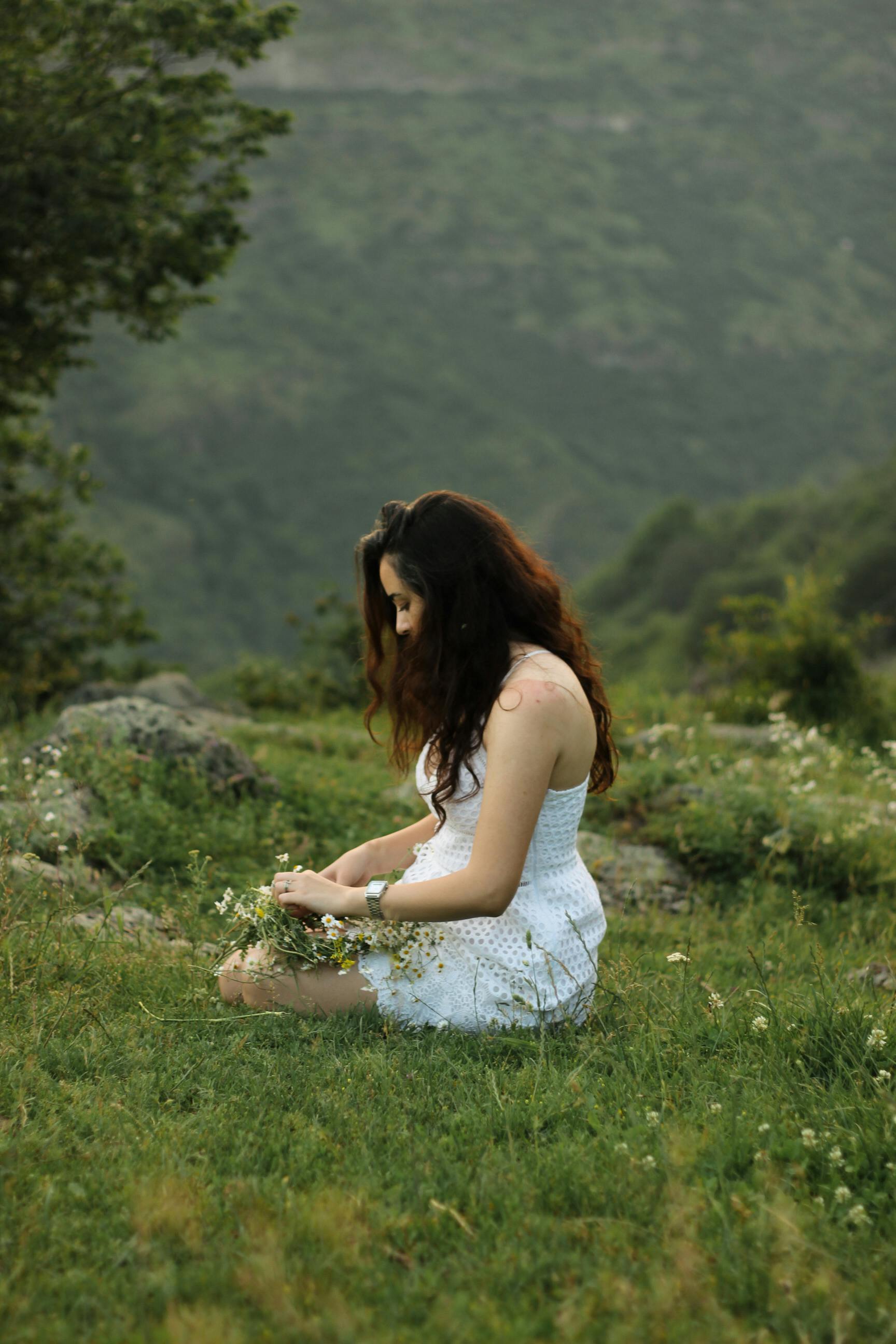 A woman in a white dress sits peacefully in a lush green meadow surrounded by hills.