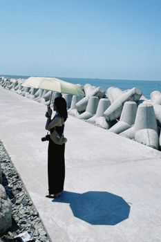 Woman with umbrella walks on a sunny concrete promenade near the sea, showcasing elegance and style.