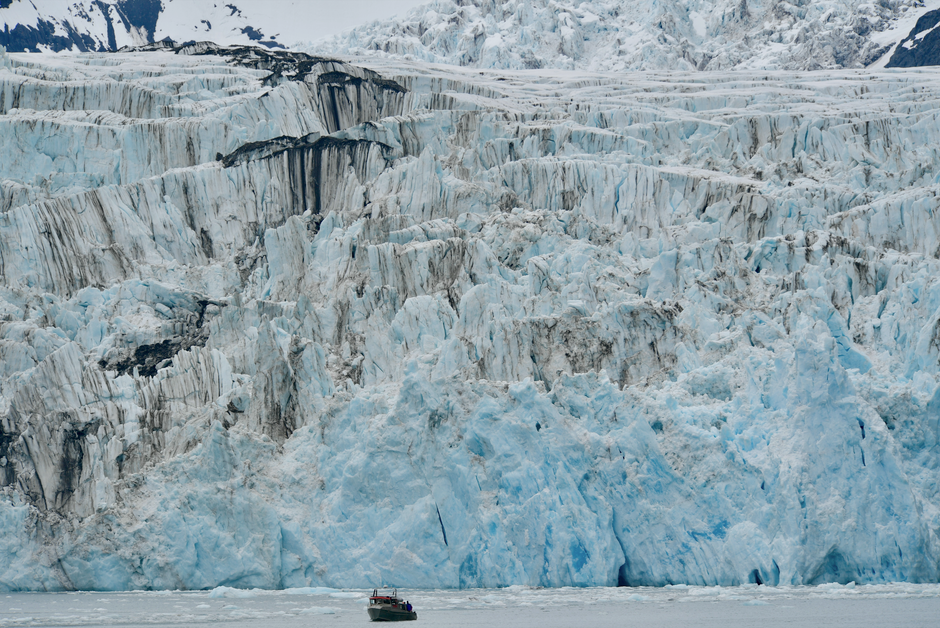 A vast glacier with icy formations and a small motorboat in the cold waters below.