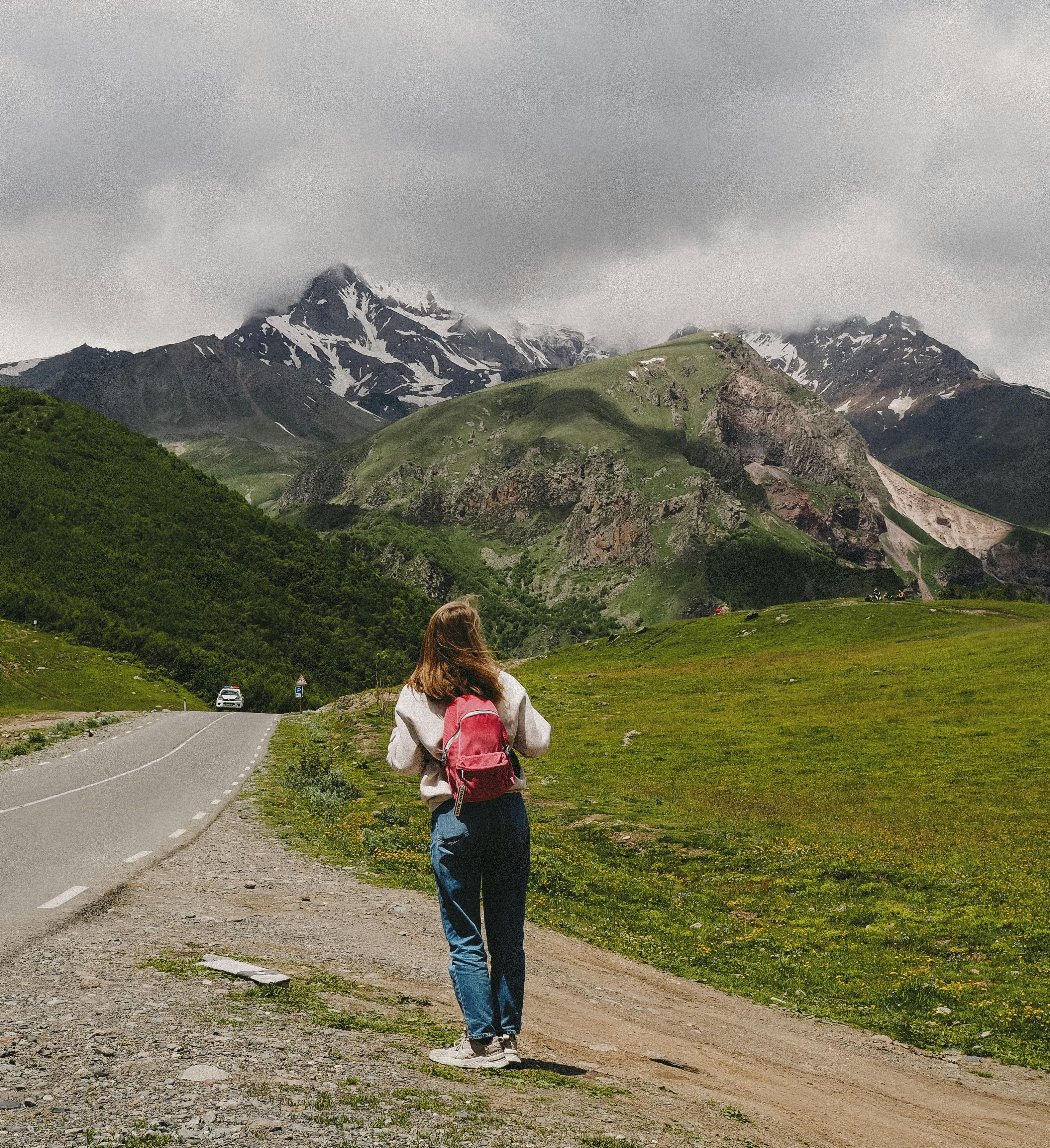 A woman with a backpack hikes beside a road with stunning mountain views.