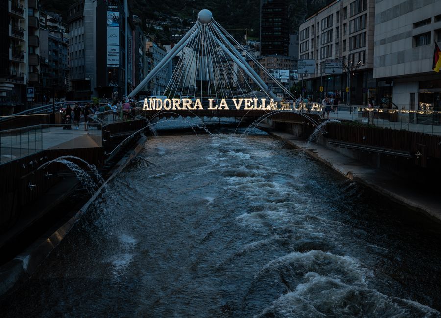Night view of the iconic Andorra la Vella bridge over the urban river, with city lights and flowing water.