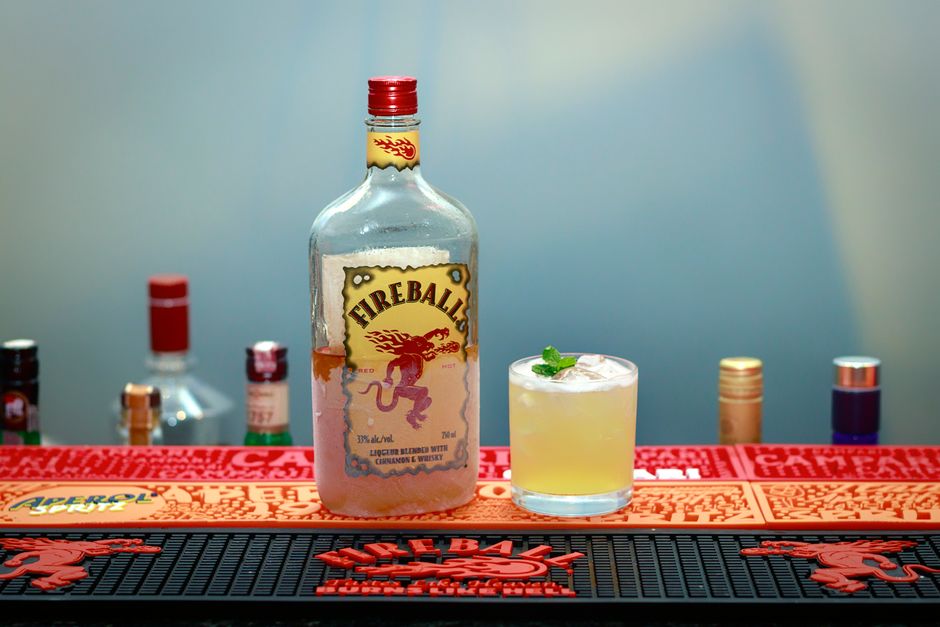 Two mugs of warm amber apple cider spiked with Fireball whiskey, each garnished with a cinnamon stick and apple slice, steam rising, surrounded by whole cloves and orange slices.