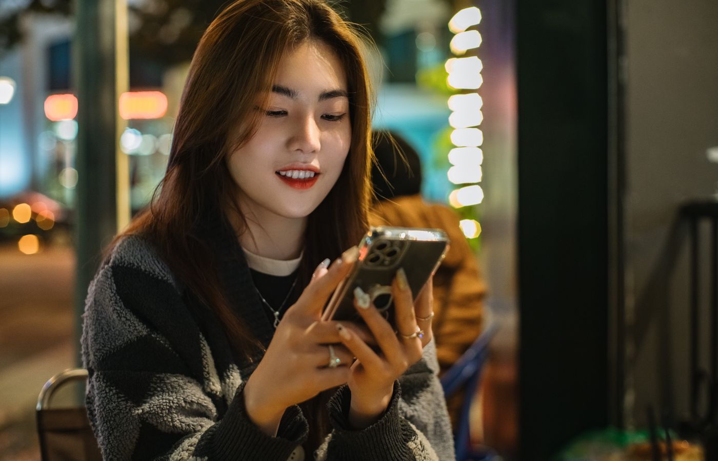 Smiling young woman sitting outdoors, texting on her smartphone during the evening.