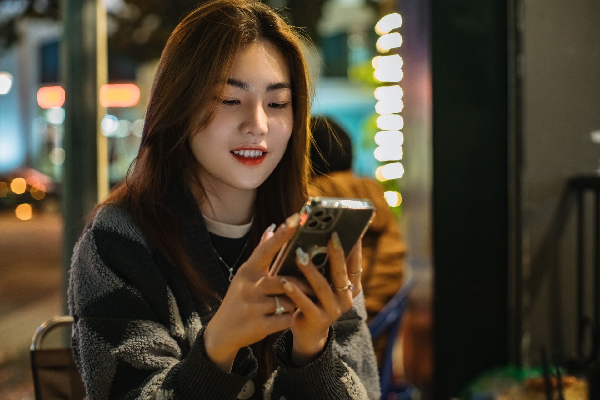 Smiling young woman sitting outdoors, texting on her smartphone during the evening.